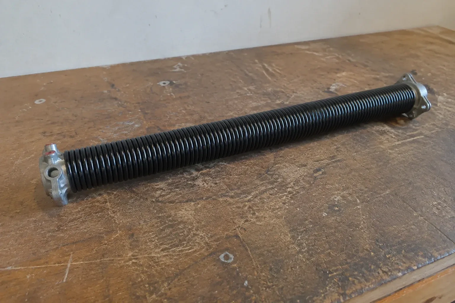 Detached black coil spring with metal end fittings on a workbench. — Torsion Spring Replacement, Brampton