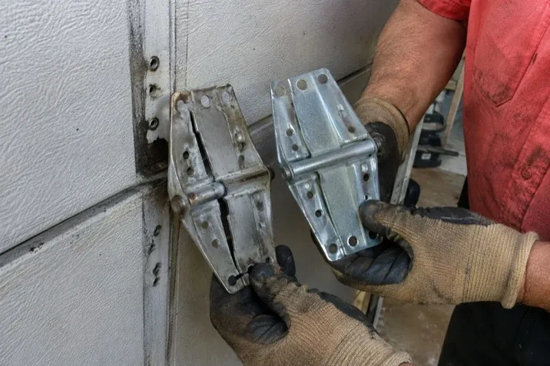Gloved hands holding two metal garage-door hinges side by side, one discolored with a visible crack and the other bright silver. — Garage Door Hinge Replacement, Brampton